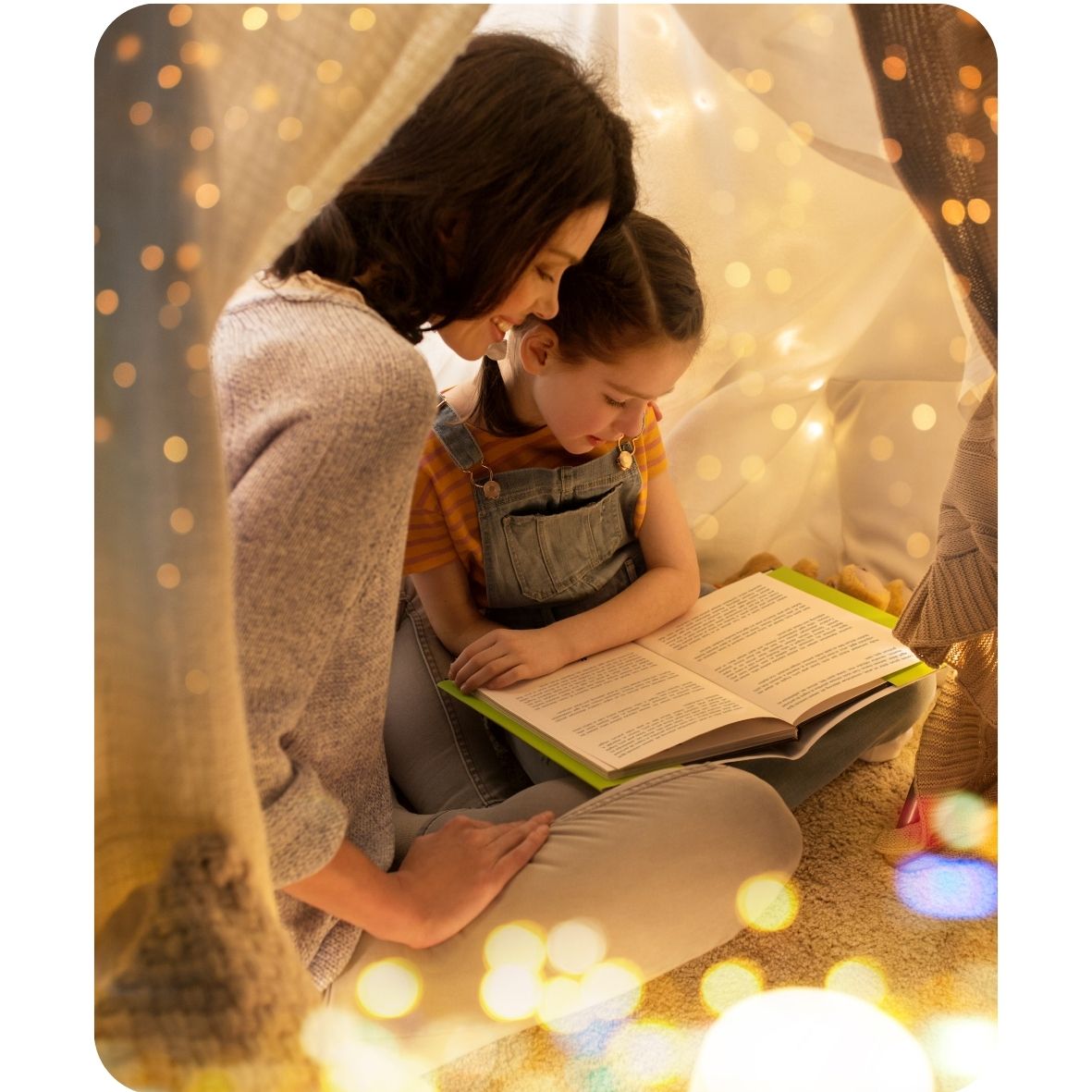Woman and child reading a book together under a blanket with string lights.