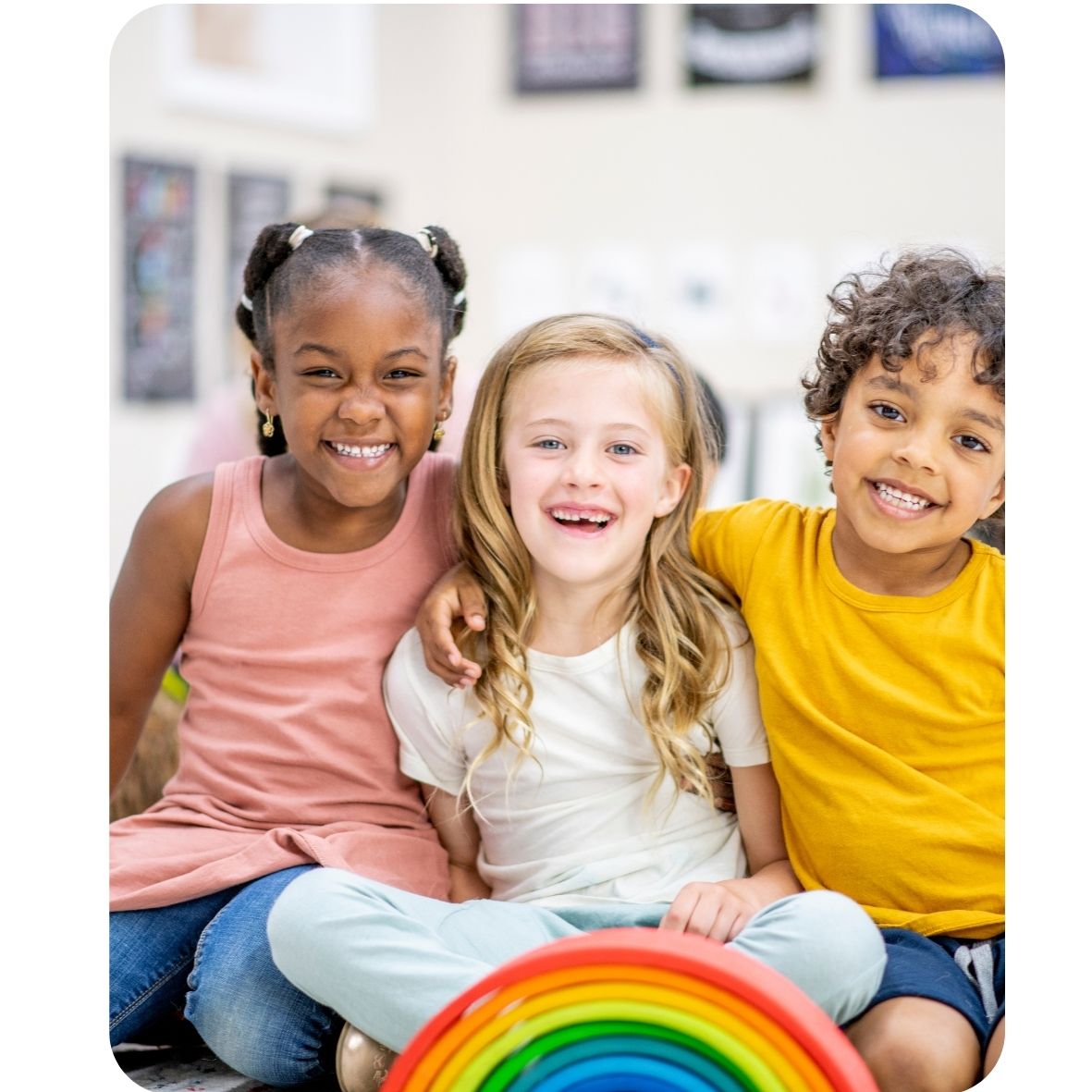 Three children sitting together with a rainbow object in a classroom setting