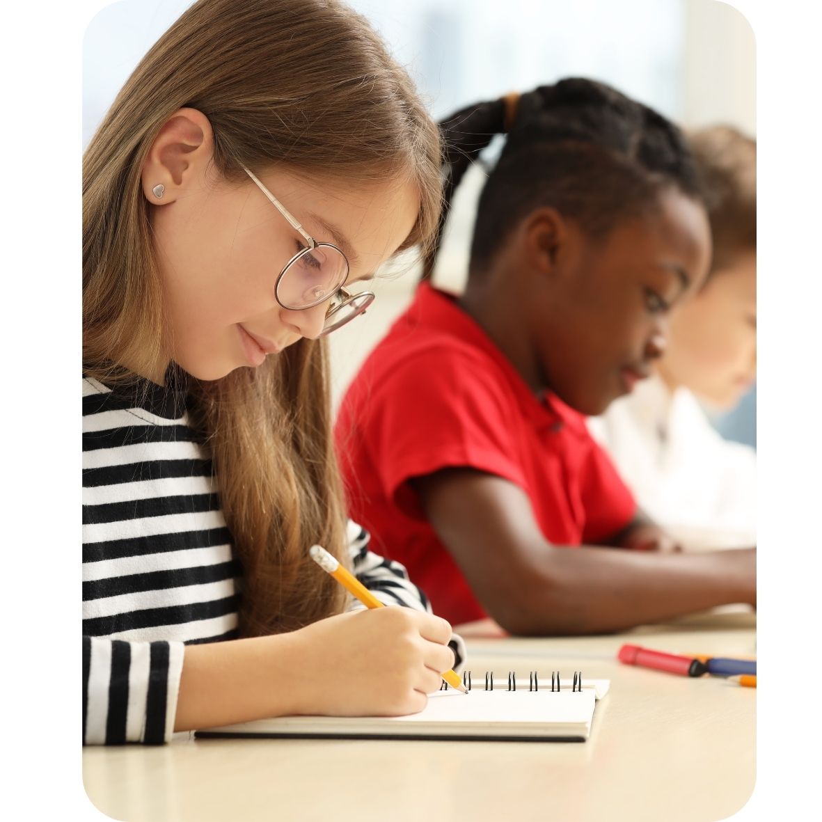 Girl with glasses writing a book with classmates in the background